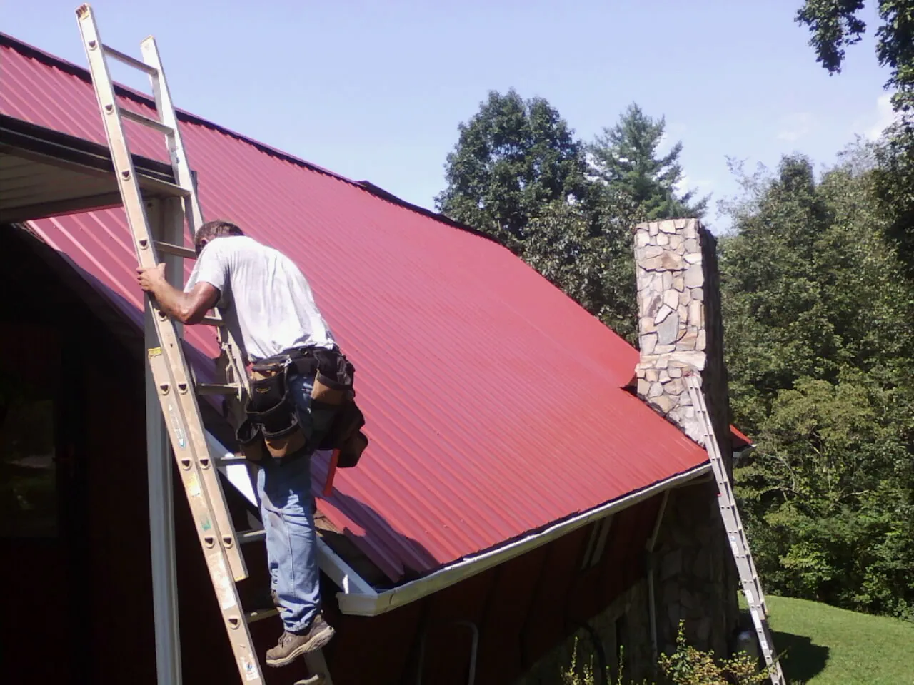 Roofer on a ladder beside a red metal roof and stone chimney — Soffit & Fascia Repair by Roofing Brothers in Yorktown Heights